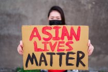 Mujer étnica en máscara y con cartel de cartón con inscripción Asian Lives Matter protestando en la calle de la ciudad y mirando a la cámara - foto de stock