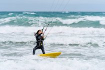 Активная спортсменка на кайтборде держит планку управления, практикуя кайтсерфинг и глядя вдаль на пенный океан — стоковое фото