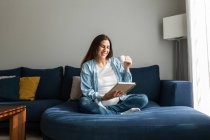 Положительная беременная женщина сидит на мягком диване с ноутбуком и пьет горячий напиток — стоковое фото