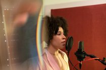 Черная певица исполняет песню против микрофона с поп-фильтром, стоя и глядя вперед в звукозаписывающей студии — стоковое фото
