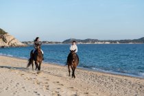 Giovane donna con fidanzato cavalcando stalloni di razza pura sulla riva sabbiosa contro l'oceano ondulato sotto il cielo blu — Foto stock