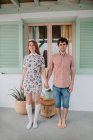 Повний вміст тіла молода пара носить повсякденне літнє вбрання, тримаючись за руки і дивлячись на камеру, стоячи біля сучасного маленького котеджу — стокове фото