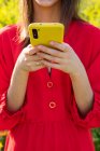 Crop female in red apparel text messaging on cellphone against blossoming plants in sunlight — Stock Photo