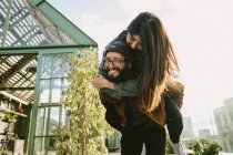 Smiling bearded boyfriend piggybacking happy girlfriend while having fun on terrace on sunny day — Stock Photo