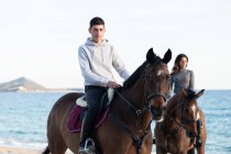 Young woman with boyfriend riding purebred stallions on sandy shore against wavy ocean under blue sky — Stock Photo