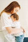 Вид сбоку молодая мама в повседневной одежде обнимает и целует маленького ребенка в светлой комнате — стоковое фото