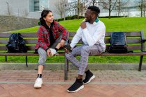 Glad multiethnic couple in stylish clothes speaking while looking at each other on city bench in daylight — Stock Photo