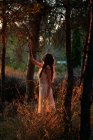 Full body of calm female in white dress standing at tree trunk in dark woods in calm sundown light - foto de stock