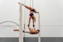 Full length sporty strong woman performing upside down split on aerial silks and lifting muscular male acrobat on hands while training together in fitness center — Stock Photo