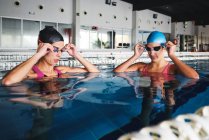 Deportivas en gorras y bañadores preparándose para entrenar en piscina con agua transparente durante el día - foto de stock
