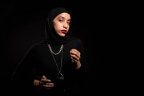 Привлекательная молодая исламская женщина в черной одежде и хиджабе нежно смотрит в камеру на черной студии — стоковое фото