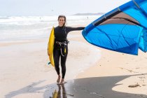 Sportlerin im Neoprenanzug mit aufblasbarem Drachen spaziert am Sandstrand und blickt gegen stürmischen Ozean — Stockfoto