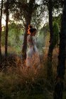 Full body of calm female in white dress standing at tree trunk in dark woods in calm sundown light — Stock Photo