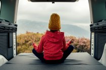 Vista trasera de una adolescente anónima sentada en furgoneta en pose de Loto y haciendo yoga mientras medita durante un viaje por las montañas - foto de stock