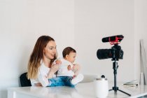 Jovem mãe com bebê bonito em voltas falando e gravando vídeo na câmera para blog pessoal enquanto sentado na mesa em casa — Fotografia de Stock