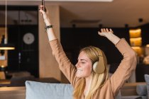Optimistic female sitting on sofa with eyes closed listening to music in headphones while enjoying songs with raised hands — Stock Photo