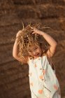 Cheerful adorable child in overalls playing with hay near straw bales in countryside — Stock Photo