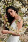 Young female with tattooed arm wearing white dress and standing in flowers of tree looking at camera — Photo de stock