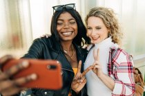 Content multiethnic female friends standing on city street and taking self shot on mobile phone at weekend — Stock Photo