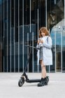 Full body of focus young African American female in blue coat browsing on smartphone while standing on sidewalk with scooter — Foto stock
