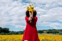 Anonymous female in hat covering face with blossoming yellow flowers in countryside field under cloudy sky — Stock Photo