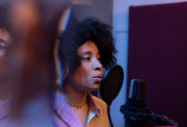 Black female singer performing song against microphone with pop filter while standing and looking forward in sound studio — Stock Photo