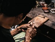 From above cropped unrecognizable concentrated ethnic elderly male jeweler in casual clothes and eyeglasses sharpening instruments while working at wooden table in traditional workshop — Stock Photo