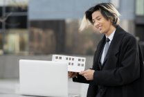 Contenido joven empresario étnico masculino con maqueta de construcción hablando en videollamada contra netbook en la ciudad - foto de stock