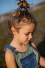 Очаровательная маленькая девочка в комбинезоне, стоящая на лугу и смотрящая вниз — стоковое фото