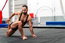 Masculino étnico musculoso de cuerpo completo en pantalones cortos deportivos que realizan Twisted Monkey Pose mientras practican yoga en un moderno gimnasio mirando a la cámara - foto de stock