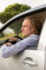 Side view of cheerful young haired male keeping hand out of car window while enjoying summer journey in nature looking away — Stock Photo