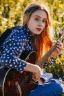 Задумчивая молодая хипстерша, сидящая на лугу в сельской местности, пишет песни на блокноте и играет на гитаре во время летнего солнца. — стоковое фото