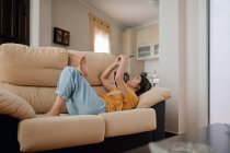 Вид збоку молодої жінки текстові повідомлення на мобільний телефон, лежачи на дивані у вітальні — стокове фото