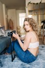 Side view of young concentrated female photographer with curly blond hair in lace bra and jeans sitting on soft carpet and taking photos on camera at home — Fotografia de Stock