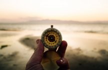 POV crop anonymous adventurer checking route with retro compass while standing on sandy beach near sea at sunset — Photo de stock