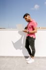 Seitenansicht fokussierter junger ethnischer Freiberufler mit dunklen Haaren im trendigen Outfit, der auf der Terrasse steht und während der Fernarbeit Laptop benutzt — Stockfoto