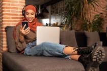 Jovem muçulmana feliz de comprimento total em roupas modestas hijab e fones de ouvido sem fio sentados no sofá acolhedor com netbook em voltas e smartphone de navegação — Fotografia de Stock