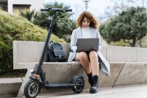 Focused young African American female in blue coat working on netbook while sitting on stone bench near scooter in city park on clear spring day — Stock Photo