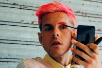 Hombre homosexual con piercings y corte de pelo moderno aplicando rímel en las pestañas con aplicador contra teléfono celular - foto de stock