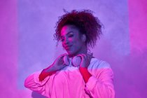 Portrait of joyful young African American female in white clothes wearing headphones on neck and looking at camera while standing in dark studio in neon lights — Stock Photo