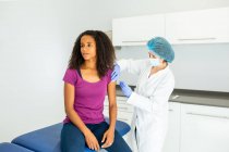 Especialista médica femenina en uniforme protector, guantes de látex y mascarilla facial vacunando a una paciente afroamericana en clínica durante el brote de coronavirus - foto de stock