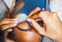 High angle of crop unrecognizable cosmetologist with tweezers applying fake eyelashes for extension on eye of ethnic client with face protective mask in salon during coronavirus pandemic — Photo de stock