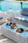 From above of plates with assorted appetizing pastries and fresh sliced watermelon served on table with cups of coffee near wicker sofa at poolside in tropical resort — Fotografia de Stock