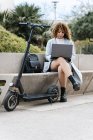 Fokussierte junge Afroamerikanerin in blauem Mantel arbeitet am Netbook, während sie an klaren Frühlingstagen auf einer steinernen Bank in der Nähe eines Motorrollers im Stadtpark sitzt — Stockfoto