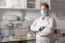 Чоловічий хімік у стерильній масці, рукавичках та захисних окулярах дивиться на камеру проти аналізатора вологи та аналітичного балансу в лабораторії канабісу — стокове фото