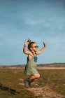 Side view of content little girl in overalls and sunglasses jumping with outstretched arms above meadow and enjoying summer on sunny day in countryside — Stock Photo