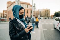 Vista lateral de la mujer musulmana en hijab y mensajes de máscara protectora en las redes sociales en el teléfono móvil mientras está de pie en la calle de la ciudad - foto de stock