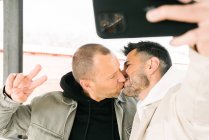 Happy young diverse homosexual guys in trendy outfits kissing and showing V sign while taking selfie on mobile phone - foto de stock
