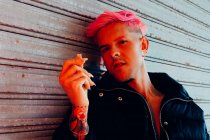 Giovane uomo omosessuale con tatuaggio e capelli rosa in capispalla alla moda guardando la fotocamera contro la parete intemperie — Foto stock