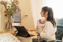 Mère adulte assise au bureau travaillant sur un ordinateur de bureau et prenant des notes dans un carnet tout en tenant un petit enfant à table pendant la journée — Photo de stock
